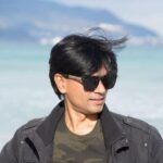 Mohammed Zubair wearing sunglasses and a black jacket stands in front of a blue sea, reminiscent of the independent journalism in India, where clarity cuts through blurred mountains of misinformation. The wind tousles their hair as they look slightly to the side.