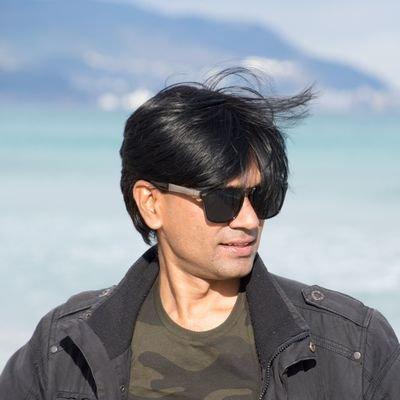 Mohammed Zubair wearing sunglasses and a black jacket stands in front of a blue sea, reminiscent of the independent journalism in India, where clarity cuts through blurred mountains of misinformation. The wind tousles their hair as they look slightly to the side.